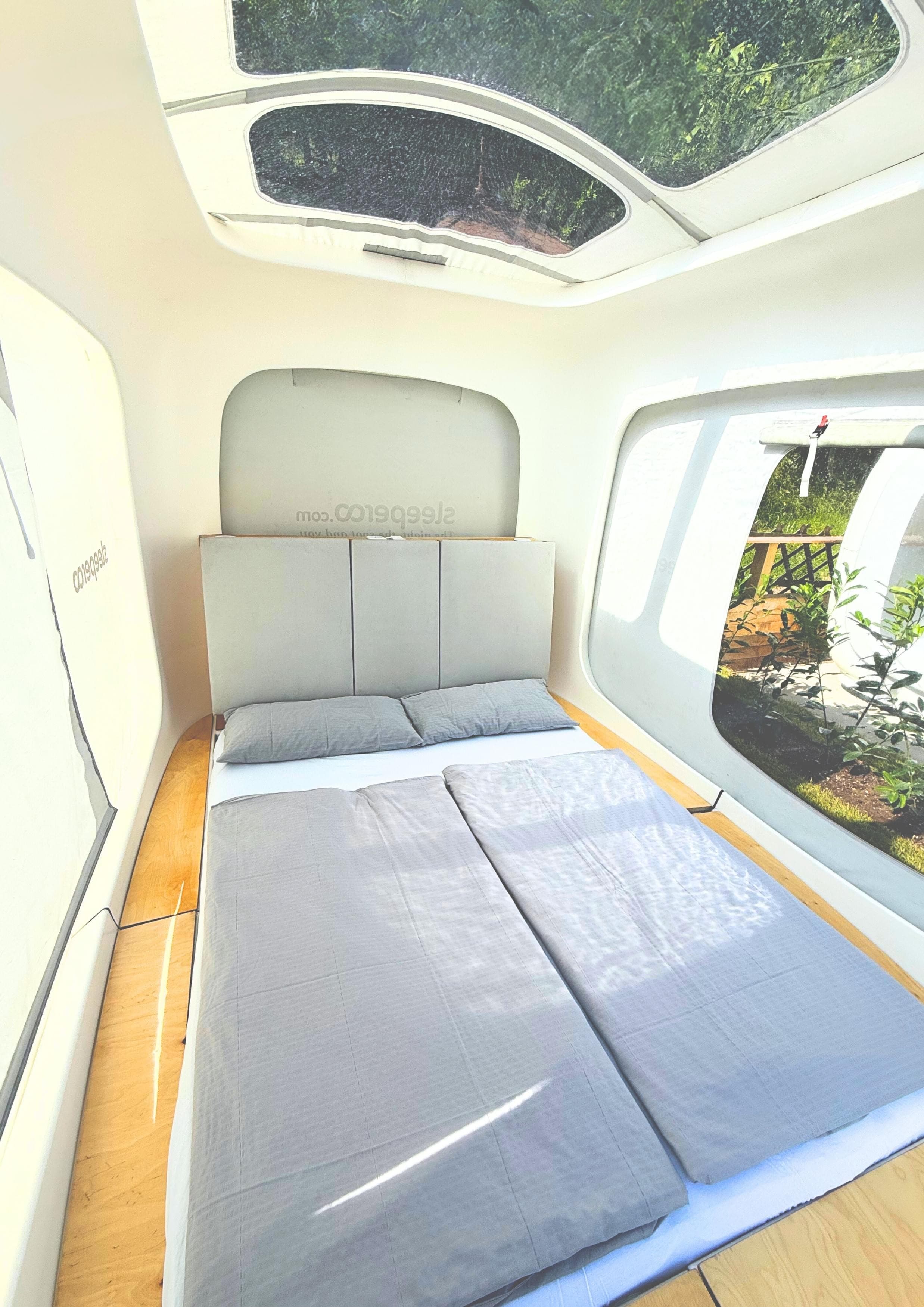 Direkt am Wallersee - Cubes - C1 - Close-up of a modern, compact sleeping pod interior with a made bed, grey bedding, and a white pillow with 'sleeperoo' logo. The headboard is grey and the surrounding walls are a soft beige fabric.